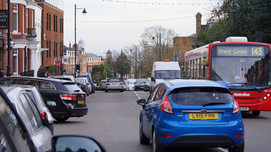 View of a busy suburban street in Brent Wembley Station and Stadium area, showing parked and moving vehicles including a blue hatchback with a UK license plate, a red Metroline bus displaying route 143 towards Brent Cross, and several cars lined along the pavement. The street is lined with brick and white-painted residential buildings, with some storefronts visible, and modern street lighting extending overhead. The scene also includes a white van extended slightly into the road, possibly involved in a home relocation or removal activity, with additional parked cars on both sides and trees in the background. Overcast skies cast diffuse natural light, and the environment suggests a typical urban setting suitable for house removals or furniture transport services provided by Brent Man and Van, as they organize loading and unloading in a residential area.