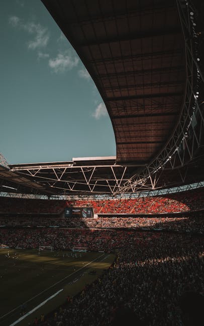 Inside a large stadium during daylight, the curved roof structure extends overhead, supported by metal trusses and beams. The seating area is filled with spectators, with a mix of red and dark-coloured seats visible in the background. The playing field features a green grass surface, marked with white lines, and a scoreboard can be seen in the distance. Despite the open roof, natural light illuminates the interior, casting shadows across the venue. This image captures the scale of the stadium and the structural elements involved in its roof design, relevant to house removals and moving logistics by Brent Man and Van, as they prepare for equipment transportation or venue setup.