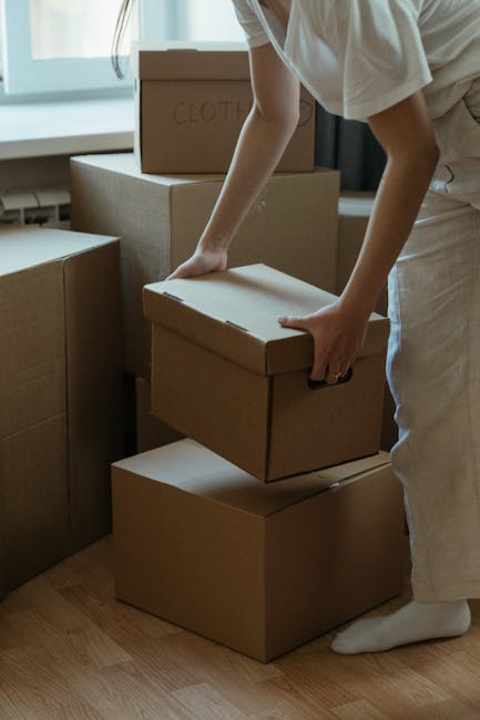 A person wearing a white t-shirt and blue jeans is kneeling inside a room filled with cardboard boxes, some stacked on the floor and others nearby. They are holding a black marker and writing on a med